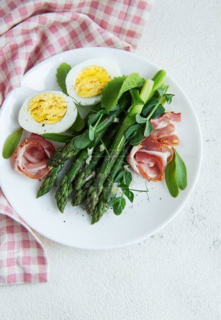A healthful salad consisting of green asparagus spears, thinly sliced prosciutto, and halves of a boiled egg, garnished with fresh microgreens leaves, is neatly arranged on a white plate. 