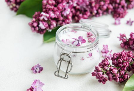 A clear glass jar with a silver clasp is filled with homemade lilac-infused sugar, adorned with delicate purple flowers. Fresh lilac blooms surround the jar, resting atop a textured white surface, hinting at a springtime culinary preparation.