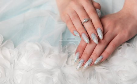 A close-up view of a persons elegantly manicured hands resting on soft, white fabric. The design features intricate blue floral art accented with glitter. A delicate silver ring with a sparkling gemstone adorns one finger, adding a touch of sophistic