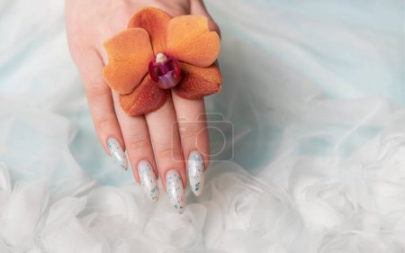 Una captura de cerca revela una mano de personas con intrincado arte floral de uñas delicadamente acunando una flor de azahar. La textura suave y ondulada que lo rodea contribuye al ambiente tranquilo y elegante de la escena.