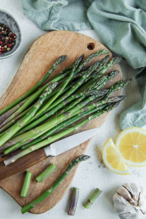 Fresh green asparagus lies on a wooden cutting board alongside a kitchen knife, with slices of lemon and garlic cloves nearby. The cutting board is on a tabletop adorned with a green cloth, and peppercorns are visible in the background, suggesting a 
