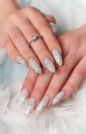 A close-up view of a persons elegantly manicured hands resting on soft, white fabric. The design features intricate blue floral art accented with glitter. A delicate silver ring with a sparkling gemstone adorns one finger, adding a touch of sophistic