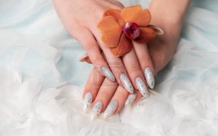 Una captura de cerca revela una mano de personas con intrincado arte floral de uñas delicadamente acunando una flor de azahar. La textura suave y ondulada que lo rodea contribuye al ambiente tranquilo y elegante de la escena.