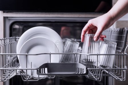Female hand loading dished, empty out or unloading dishwasher with utensils. Kitchen appliances, lifestyle view. Woman puts a plate in the dishwasher or takes from it. Housewife does her housework 