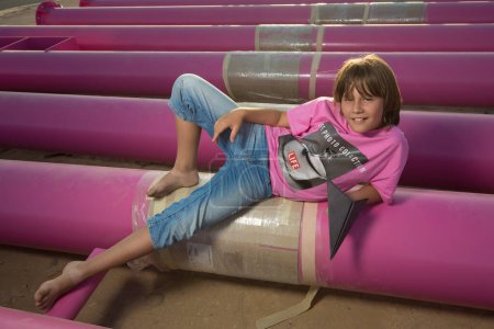 Photo for A cute caucasian boy is playing with a paper plane in a pink stylish printed t-shirt among pink iron pipes on an industrial background. Fashion photography - Royalty Free Image