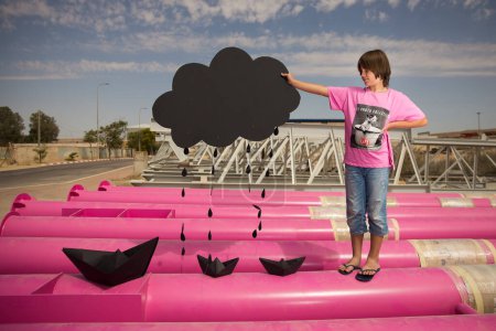 Téléchargez les photos : Un mignon enfant dans un t-shirt rose avec un imprimé chat joue parmi les tuyaux roses avec des nuages noirs, de la pluie, des bateaux et des avions en papier contre le ciel bleu dans un endroit industriel. photoshoot style mode - en image libre de droit