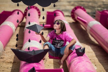 Foto de Un niño lindo en una camiseta rosa con un gato print juega entre las tuberías de color rosa con nubes negras, lluvia, barcos y aviones de papel contra el cielo azul en un lugar industrial. sesión de fotos de estilo de moda - Imagen libre de derechos