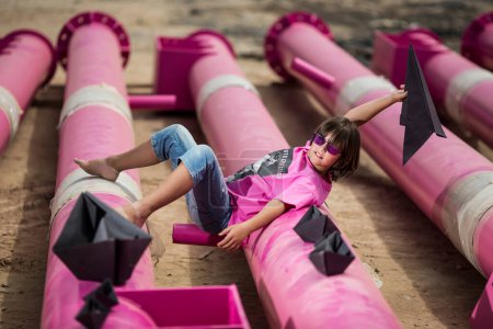 Foto de Un niño lindo en una camiseta rosa con un gato print juega entre las tuberías de color rosa con nubes negras, lluvia, barcos y aviones de papel contra el cielo azul en un lugar industrial. sesión de fotos de estilo de moda - Imagen libre de derechos