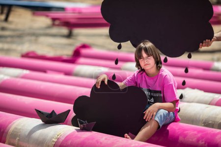 Téléchargez les photos : Un mignon enfant dans un t-shirt rose avec un imprimé chat joue parmi les tuyaux roses avec des nuages noirs, de la pluie, des bateaux et des avions en papier contre le ciel bleu dans un endroit industriel. photoshoot style mode - en image libre de droit