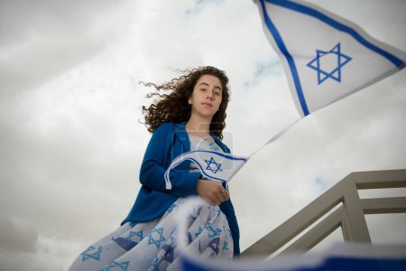 Hermosa chica judía de cabello oscuro o mujer con símbolos judíos de Israel vestidos con los colores de la bandera israelí contra el cielo nublado afuera, concepto del Día de la Independencia