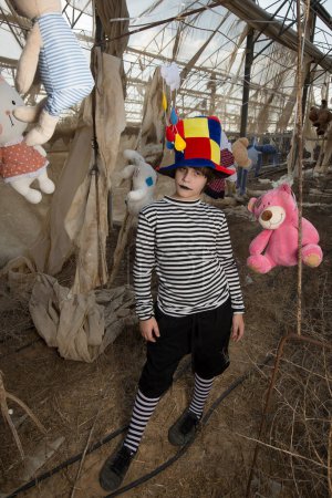 Téléchargez les photos : Un garçon avec un sourire sinistre dans un costume de clown joue et s'amuse dans une serre abandonnée avec un perroquet dans une cage et des jouets en peluche - en image libre de droit