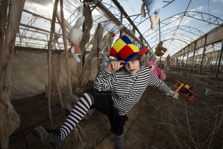 Téléchargez les photos : Un garçon avec un sourire sinistre dans un costume de clown joue et s'amuse dans une serre abandonnée avec un perroquet dans une cage et des jouets en peluche, contre un ciel nuageux bleu - en image libre de droit