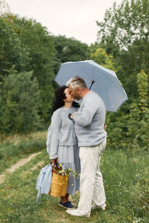 Photo for Close up romantic couple standing in autumn park under umbrella in daytime. Man and woman wearing blue sweaters. Man and woman kissing. - Royalty Free Image