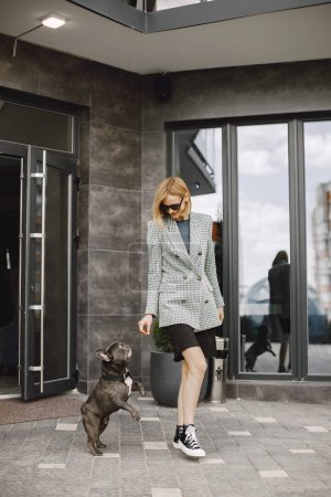Foto de Mujer joven parada cerca de la cafetería al aire libre con bulldog francés negro. Chica con gafas de sol negras, pantalones cortos y chaqueta gris. Mujer jugando con un perro. - Imagen libre de derechos