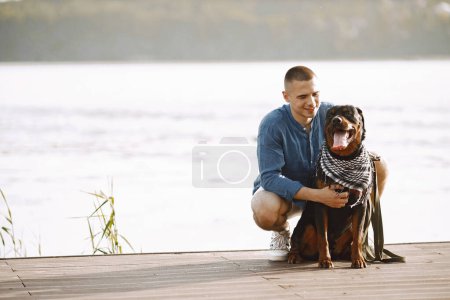Téléchargez les photos : Handsome young male in casual outfit petting cute dog while sitting near the lake. Boy wearing blue shirt and white jeans shorts. Dog has a white and black scarf on his neck. - en image libre de droit