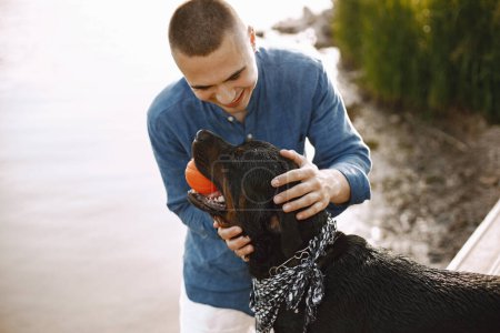 Téléchargez les photos : Handsome young male in casual outfit playing with cute dog while standing near the lake. Boy wearing blue shirt and white jeans shorts. Dog has a white and black scarf on his neck. - en image libre de droit