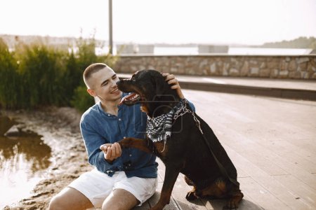 Téléchargez les photos : Handsome young male in casual outfit playing with cute dog while sitting near the lake. Boy wearing blue shirt and white jeans shorts. Dog has a white and black scarf on his neck. - en image libre de droit