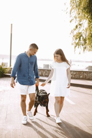 Téléchargez les photos : Romantic couple boyfriend and girlfriend walking with rotweiller dog near the lake together. Man and woman looking at each other. Man wearing blue shirt and woman white dress. - en image libre de droit