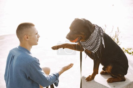 Téléchargez les photos : Handsome young male in casual outfit playing with cute dog with orange ball while sitting near the lake. Dog gives a paw. Dog has a white and black scarf on his neck. - en image libre de droit