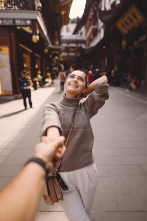 Foto de Recién casados mostrando afecto y tomados de la mano en Shanghai cerca de Yuyuan. marido y mujer tomados de la mano en la calle por un mercado y pagoda. Chica morena feliz sonriendo grande para la cámara. - Imagen libre de derechos