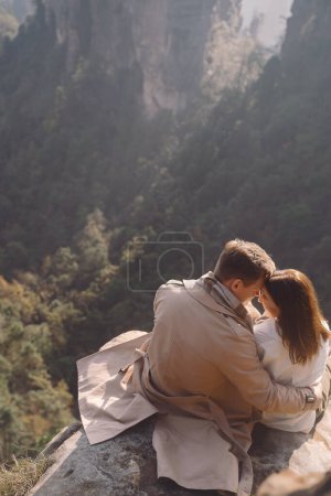 Photo for Newly enageged couple hugging and holding hands as they are sitting on a rock at zhangjiajie national forest park in China - Royalty Free Image