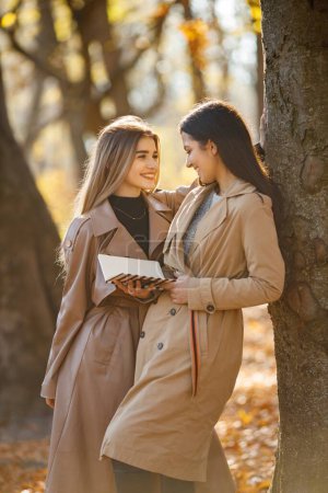 Photo for Two beautiful female friends spending time together. Two young smiling sisters talking and standing near tree with a book. Brunette and blonde girls wearing coats. - Royalty Free Image