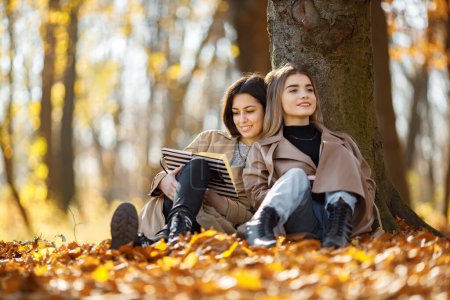 Photo for Two beautiful female friends spending time together. Two young smiling sisters sitting near tree and reading a book. Brunette and blonde girls wearing coats. - Royalty Free Image