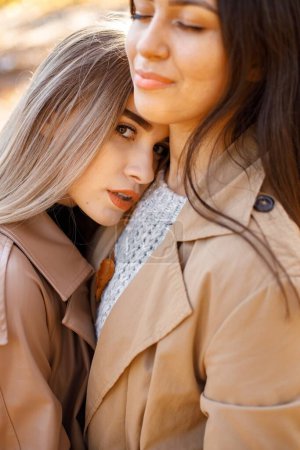 Photo for Two beautiful female friends spending time together. Two young smiling sisters walking in autumn park and hugging. Brunette and blonde girls wearing coats. - Royalty Free Image
