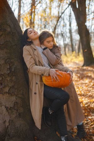 Photo for Two beautiful female friends spending time together. Two young smiling sisters talking and standing near tree with a pumpkin. Brunette and blonde girls wearing coats. - Royalty Free Image