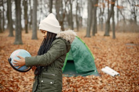 Photo for One little black girl in tent camping in the forest. Girl is holding a globe in her hands. Black girl wearing khaki coat and beige hat. - Royalty Free Image