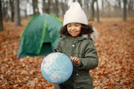 Photo for One little black girl in tent camping in the forest. Girl is holding a globe in her hands. Black girl wearing khaki coat and beige hat. - Royalty Free Image