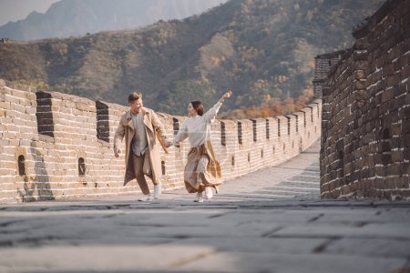 Foto de Hermosa pareja joven corriendo y saltando en la Gran Muralla de China. Pareja recién casada en su honemoon a la Gran Muralla cerca de Beijing China. Elegante pareja explorando una de las maravillas del mundo - Imagen libre de derechos