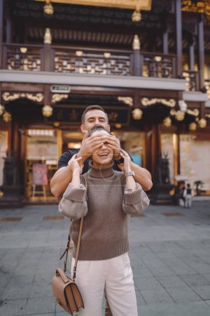 Foto de Recién casados mostrando afecto en Shanghai cerca de Yuyuan. Pareja tomar un descanso para algunos abrazos durante su visita a China. marido y mujer tomados de la mano en la calle por un mercado y pagoda - Imagen libre de derechos