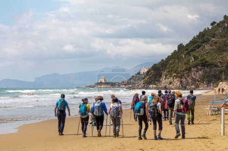 Foto de Sperlonga (LT), Italia - 25 de abril de 2022: Un grupo de excursionistas camina por la playa soleada que conduce al característico pueblo de Sperlonga. - Imagen libre de derechos