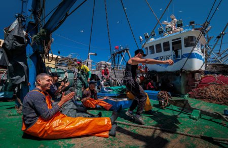 Téléchargez les photos : Essaouira, Maroc - 2 août 2023 : Un bateau de pêche ancré au quai du port avec son équipage, des hommes se relaxant, plaisantant et dansant après une longue nuit passée à la pêche en mer. - en image libre de droit