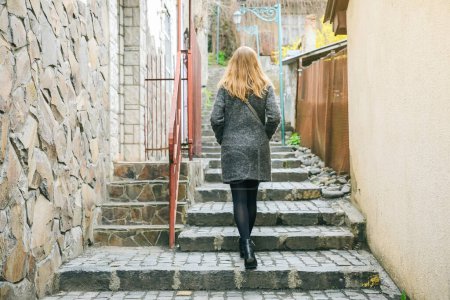 Girl is walking in the narrow street with stone pavement. Stylish woman. Medieval lantern. Stairs in the city.