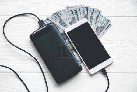 Photo for Black power bank with usb and smartphone on wooden background. Dollar notes lying nearby. Modern information technology photo. - Royalty Free Image