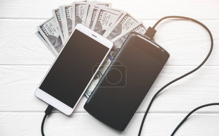 Photo for Black power bank with usb and smartphone on wooden background. Dollar notes lying nearby. Modern information technology photo. - Royalty Free Image