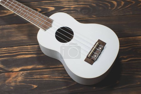 Téléchargez les photos : Guitare ukulélé en bois de couleur blanche sur le fond brun en bois. Guitare à quatre cordes hawaïenne. Instrument de musique. - en image libre de droit