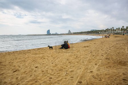 Foto de Una pareja está sentada en la playa de otoño. El perro camina sobre la arena. Foto de la atmósfera. Barceloneta. Hombre y mujer enamorados. - Imagen libre de derechos