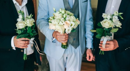 Groom in light blue suit is holding wedding bouquet. Best man friends are standing nearby.  Elegant photo.