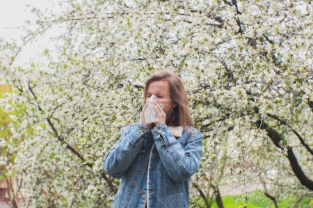 Photo for Woman sneezing into a handkerchief near a tree full of blossoms. She is suffering from seasonal spring allergy. - Royalty Free Image