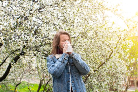 Photo for Woman sneezing into a handkerchief near a tree full of blossoms. She is suffering from seasonal spring allergy. - Royalty Free Image
