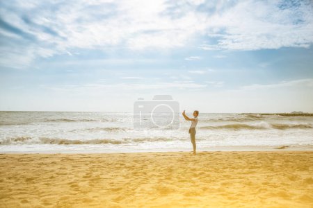 Hipster girl is taking selfie at the beach. Big waves at the ocean side. Coastline in the clouds and sun. Summer vacation. Travel concept.