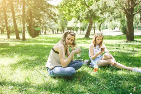 Zwei Hipster-Mädchen sitzen mit Sommercocktails im Gras. Schwestern schauen auf ihre Smartphones. Kommunikation in sozialen Netzwerken. Kalte alkoholfreie Getränke mit Eis to go. Mojito und Limonade