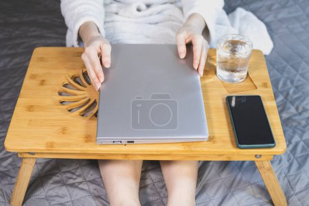 Mujer está trabajando con el ordenador portátil, smartphone y vaso de agua en la cama. Moderno portátil delgado gris en la almohadilla de enfriamiento de madera para portátil. Enfriador portátil para computadora, ventilador USB.