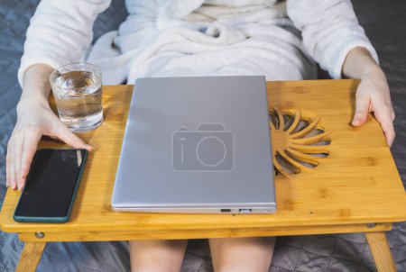 Femme travaille avec ordinateur portable, smartphone et verre d'eau sur le lit. Ordinateur portable mince gris moderne sur le coussin de refroidissement en bois pour ordinateur portable. Refroidisseur pour ordinateur portable, ventilateur USB.