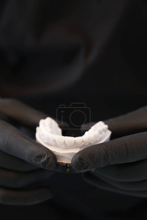 Foto de Dentista con guantes negros sosteniendo prótesis dental blanco.Cerrar prótesis dental blanca sobre fondo negro. - Imagen libre de derechos