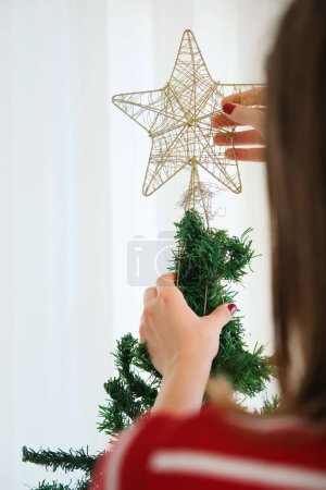 Foto de Vertical joven morena mujer en un suéter rojo pone un adorno estrella de oro en el árbol de Navidad verde.Año nuevo emoción en casa. - Imagen libre de derechos