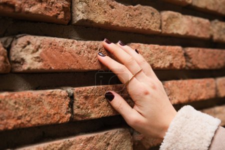Foto de Cerca de las manos de la mujer joven con esmalte de uñas de color burdeos tocar pared de baldosas industriales.Las manos de la mujer joven y la pared de baldosas rojas en el fondo. - Imagen libre de derechos
